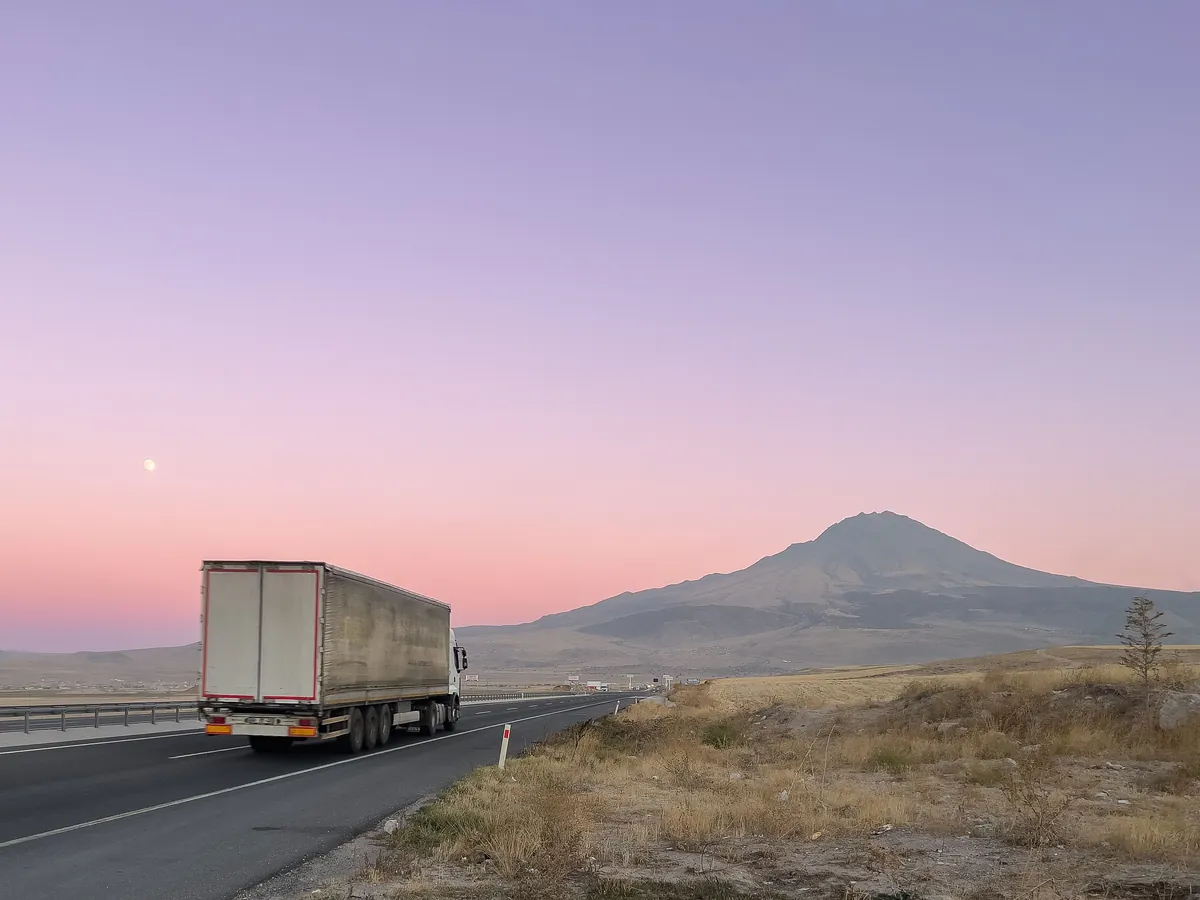 A semi-trailer truck driving along a highway towards a mountain beneath a pink dusk sky.